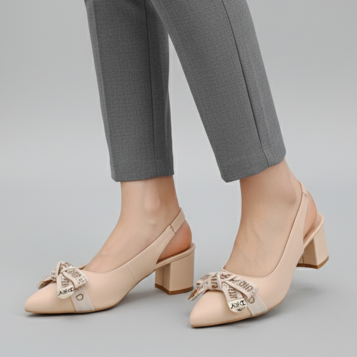 Beige high-heeled shoes with decorative bows worn with gray pants on a light gray background