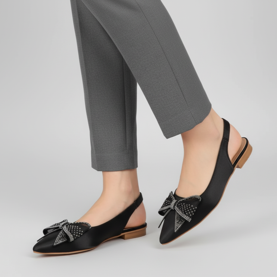 Black flat shoes with bow details worn with gray pants on a light gray background