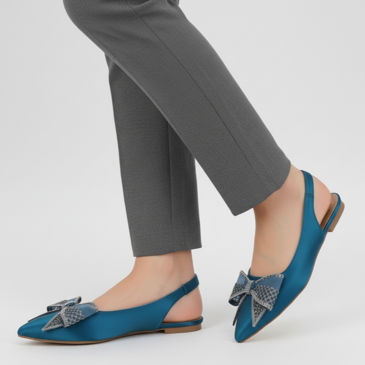 Blue flat shoes with decorative bows worn with gray pants on a white background