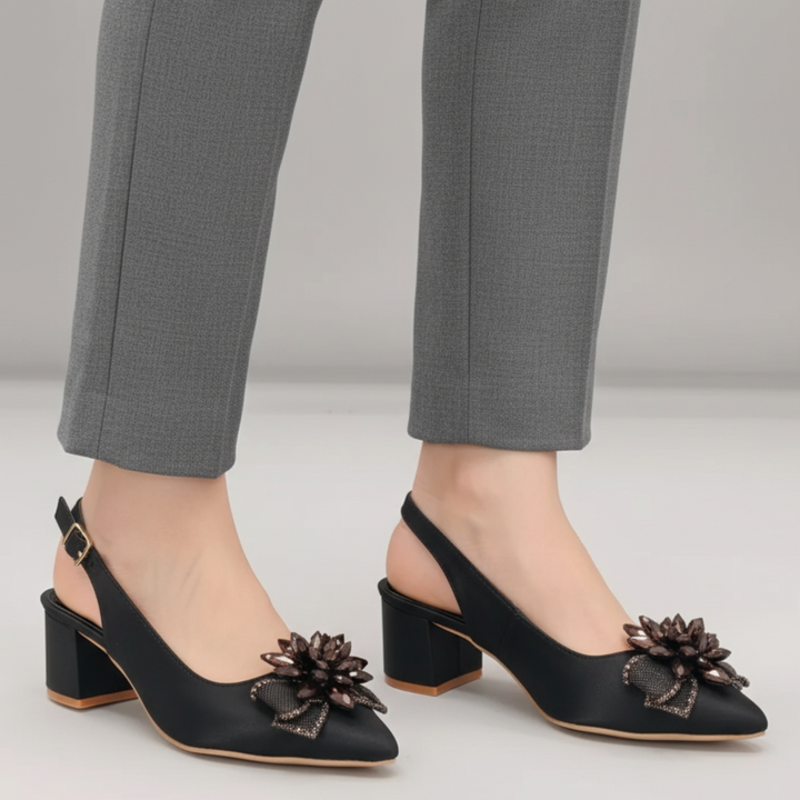 Black high-heeled shoes with floral embellishments worn with gray pants on a light gray background