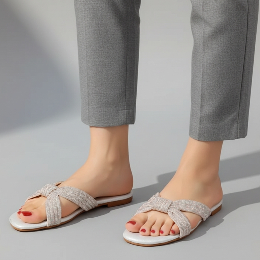 White sandals with decorative straps worn by a person in gray pants on a light gray background