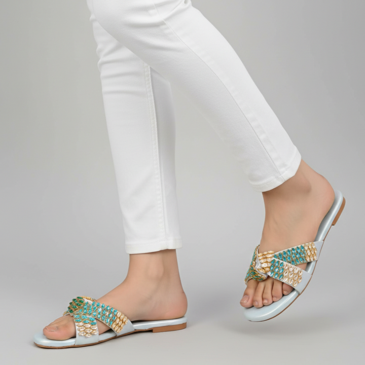 White sandals with gold and turquoise embellishments worn with white pants on a gray background
