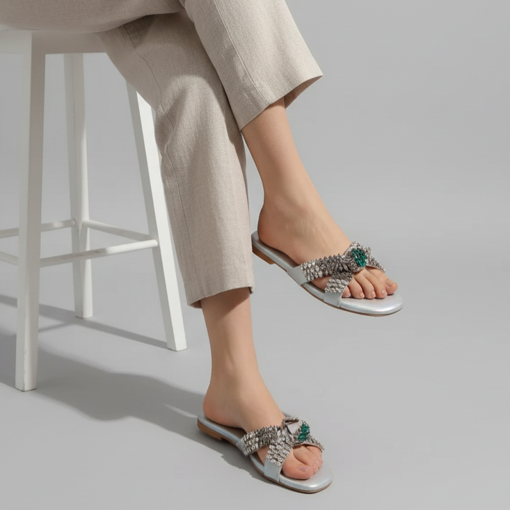 Person wearing beige pants and embellished sandals sitting on a white stool against a gray background