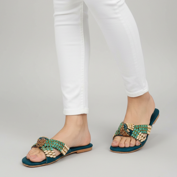 Teal sandals with gold embellishments worn with white pants on a gray background