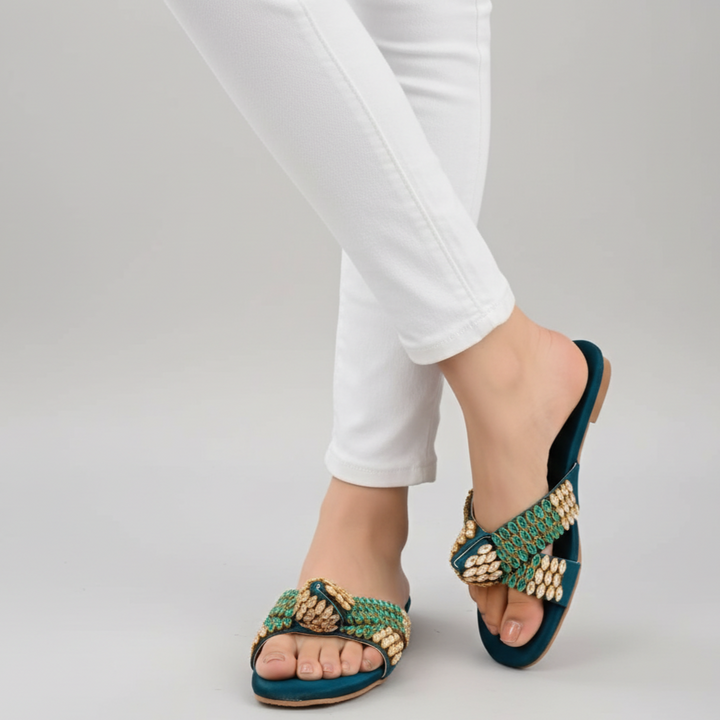 Teal sandals with gold accents worn with white pants on a light gray background