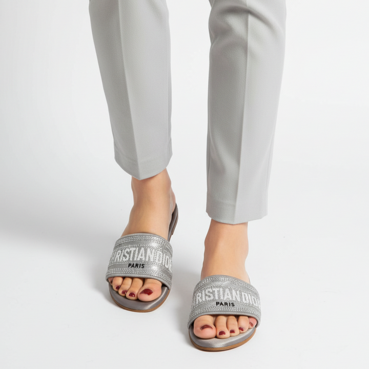 Silver sandals with 'Christian Dior' branding worn with light gray pants on a white background