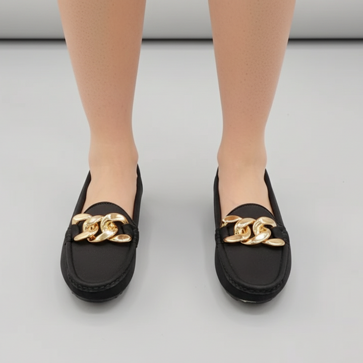 Black loafers with gold chain detail worn by a person on a light gray background
