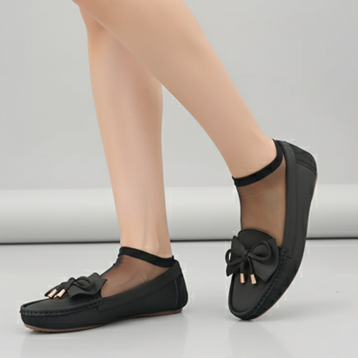 Black flat shoes with bow details worn by a person on a light gray background