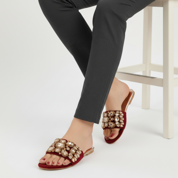 Red sandals with gold embellishments worn by a person sitting on a white stool.