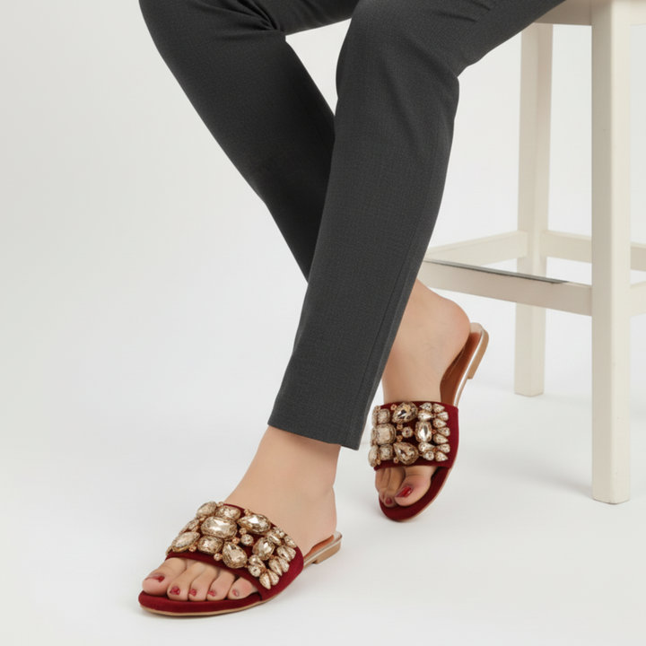 Red sandals with gold embellishments worn by a person sitting on a white stool.