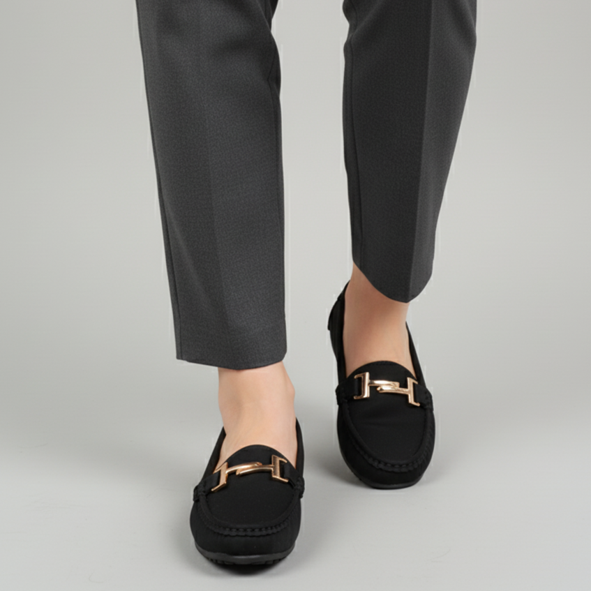 Black loafers with gold buckles worn with black pants on a light gray background