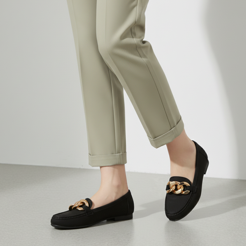 Black loafers with gold accents worn with beige pants on a light gray background