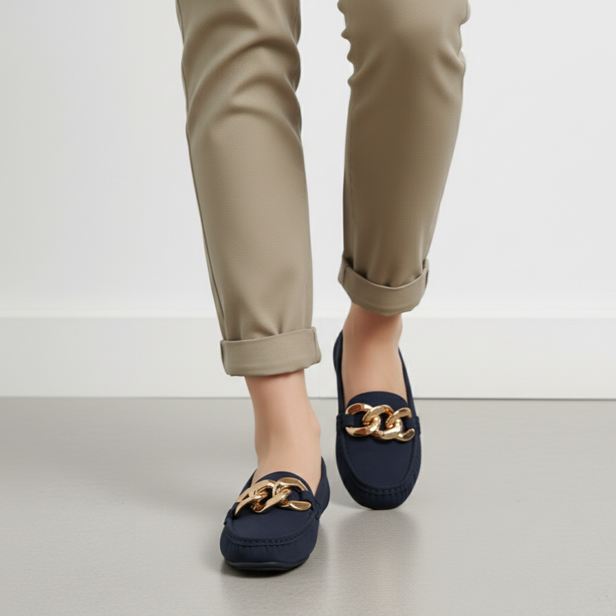 Navy loafers with gold chain detail worn with beige pants on a light gray background