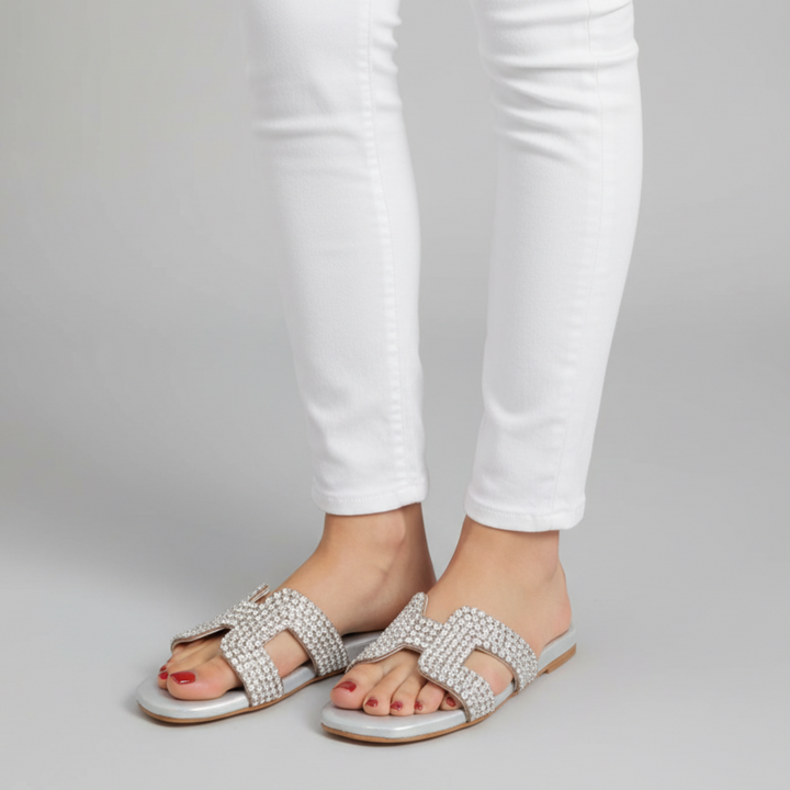 Silver sandals with gemstones worn with white pants on a gray background